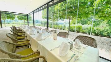 Una larga mesa de comedor preparada para una comida, con sillas de mimbre, manteles blancos y vistas al exterior.