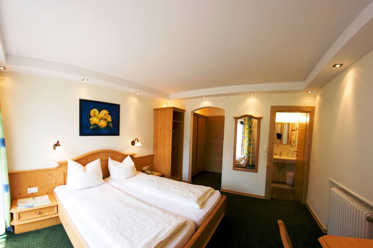 A hotel room with a large bed, a wooden wardrobe, a bathroom with a mirror, and a painting on the wall.