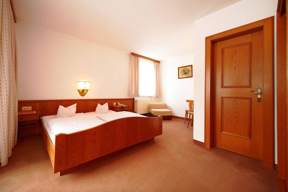 A hotel room with a wooden bed, a bedside table, a lamp, a chair, a desk, a window with curtains, and a door.