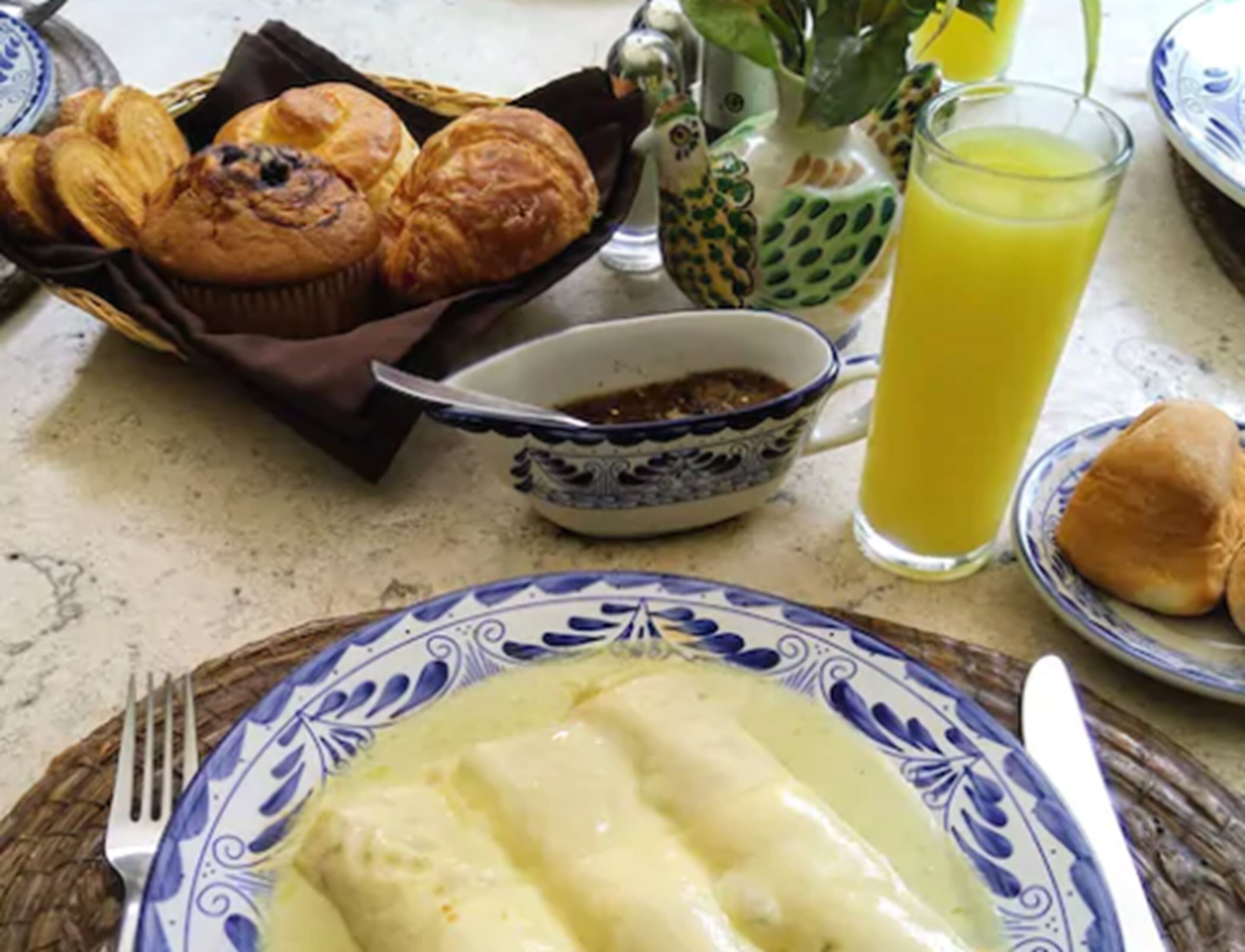 A table with a plate of enchiladas, a basket of muffins, a pineapple, a glass of orange juice, and a bowl of salsa.