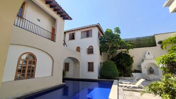A pool area with a clear blue pool, a white building with multiple windows and balconies, and a small statue in the corner.