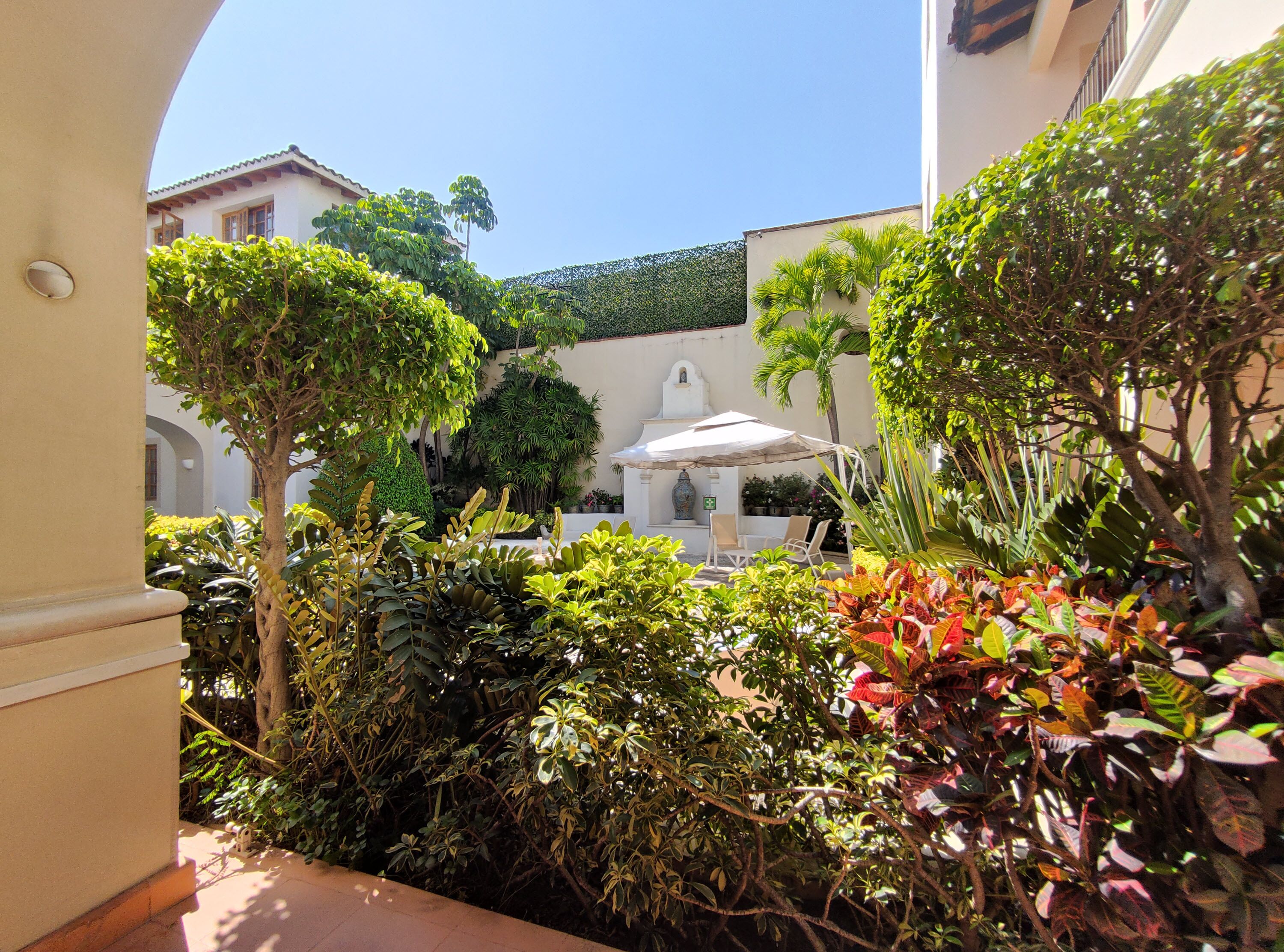 A courtyard with a white gazebo, lush greenery, and a white building with a tiled roof.