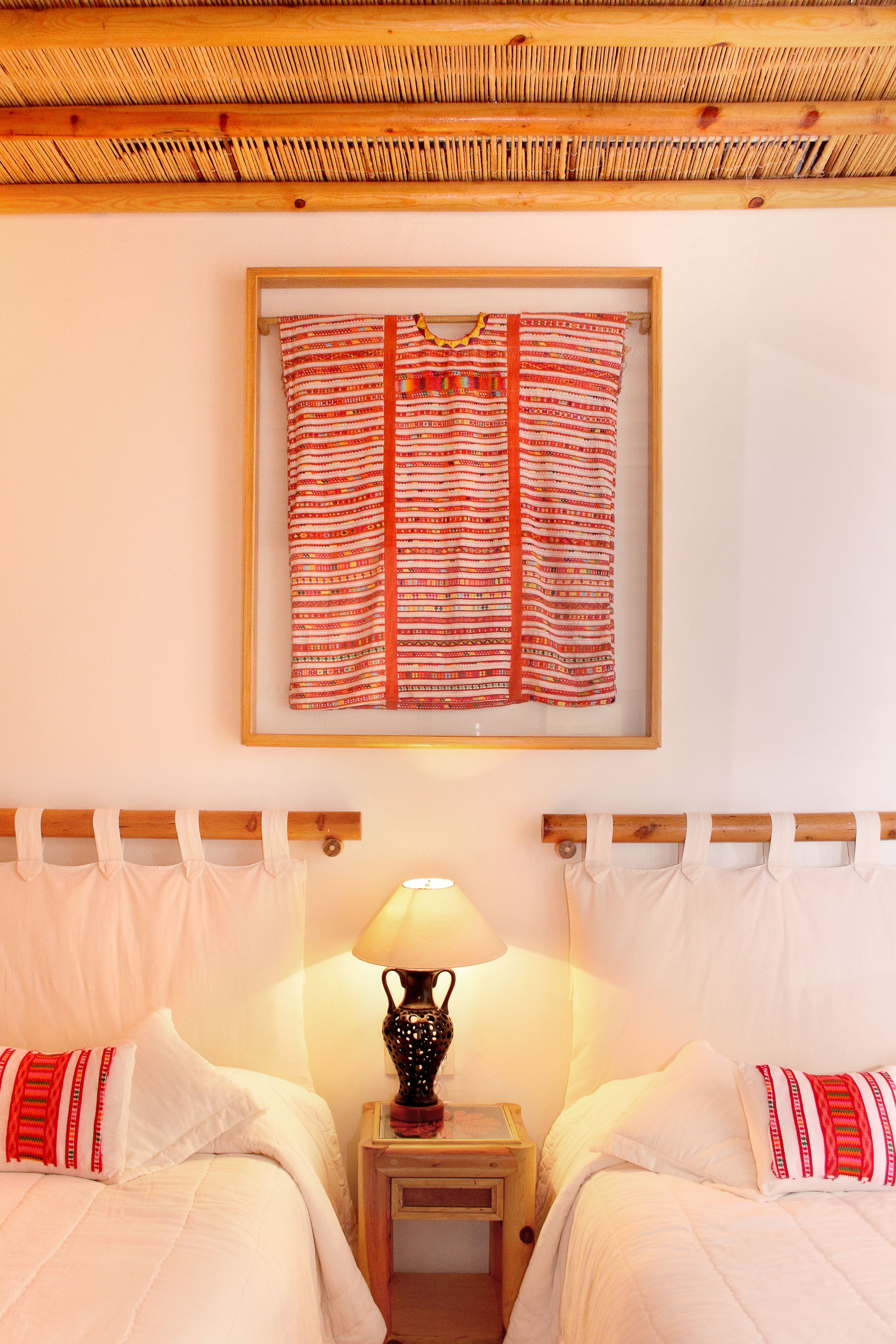 Een hotelkamer met twee bedden, een lijst met een textielkunstwerk, een bedlamp en een houten plafond.