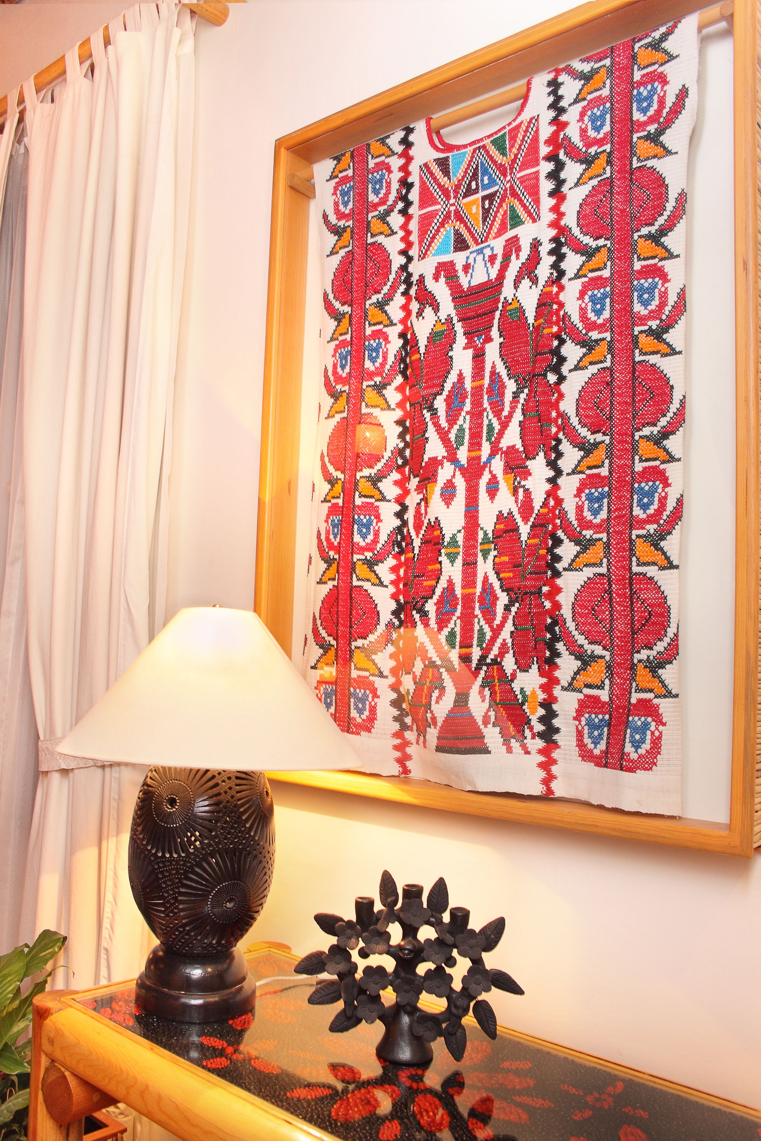 A room with a framed textile artwork on the wall, a table with a lamp and decorative items, and a window with curtains.