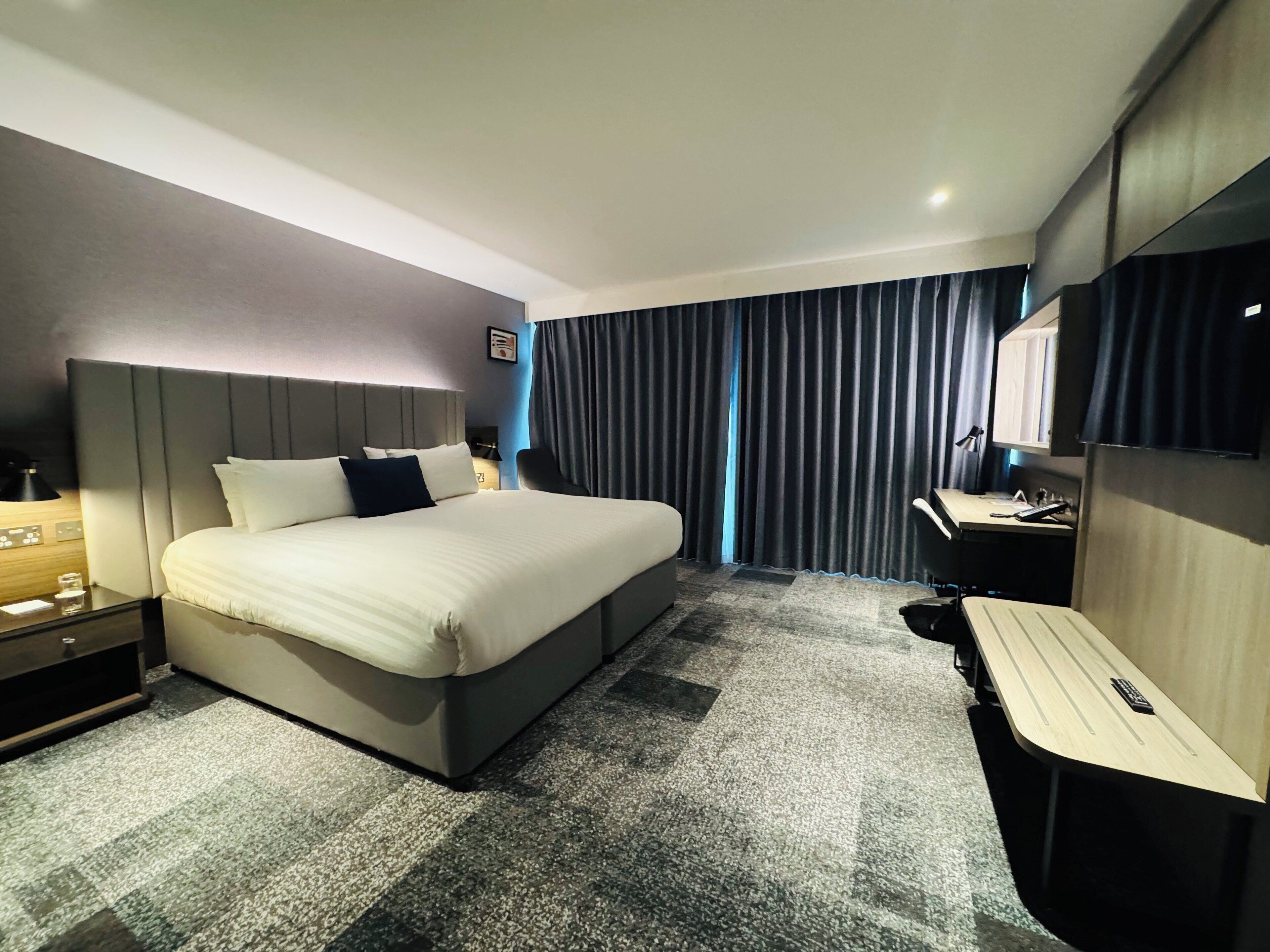 A modern hotel room with a large bed, a desk, a chair, a small table, a mirror, and a window with curtains.