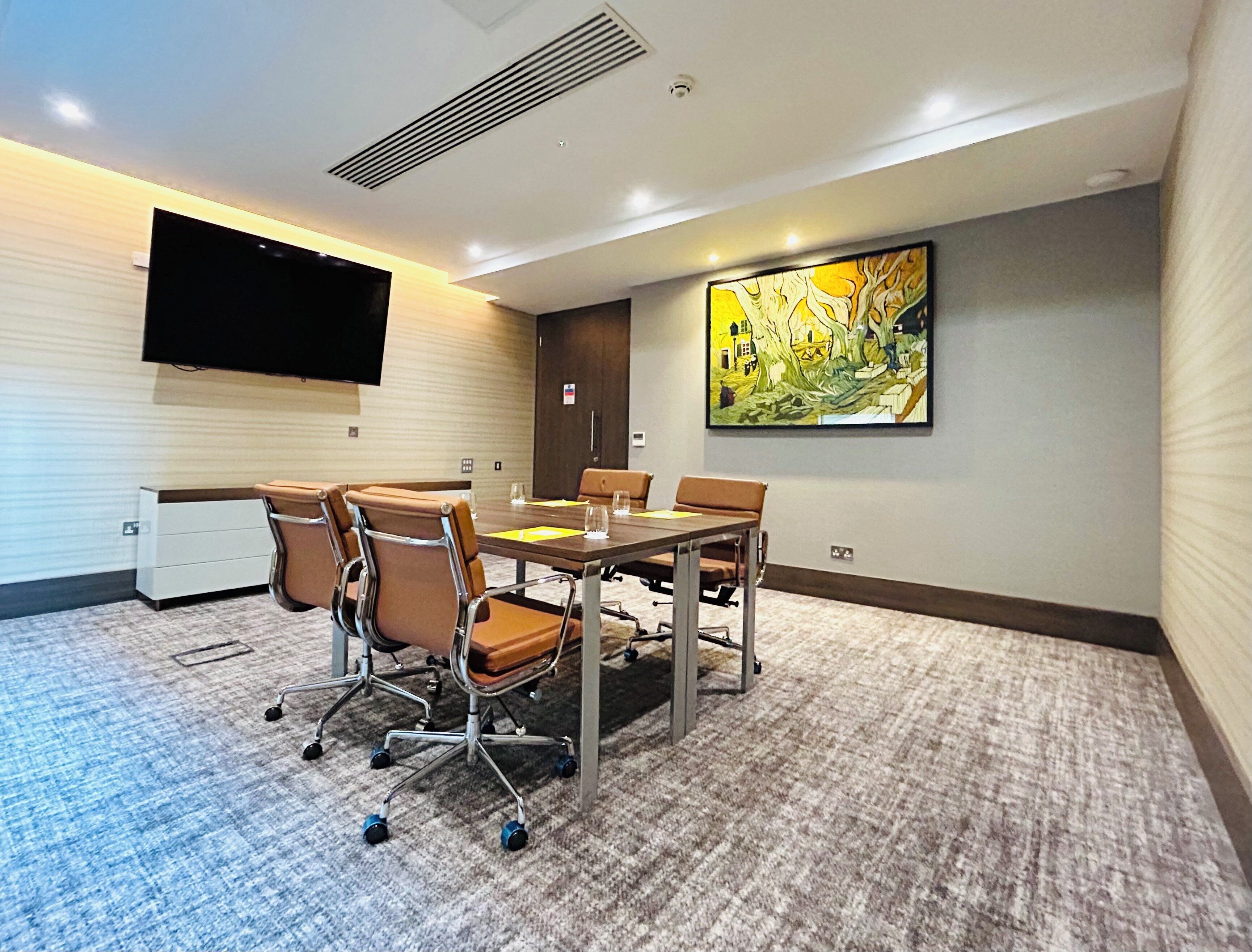 A conference room with a large table, four chairs, a flat-screen TV mounted on the wall, and a painting on the wall.