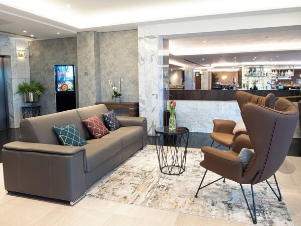A modern hotel lobby with a sofa, armchairs, a small table, and a bar area in the background.
