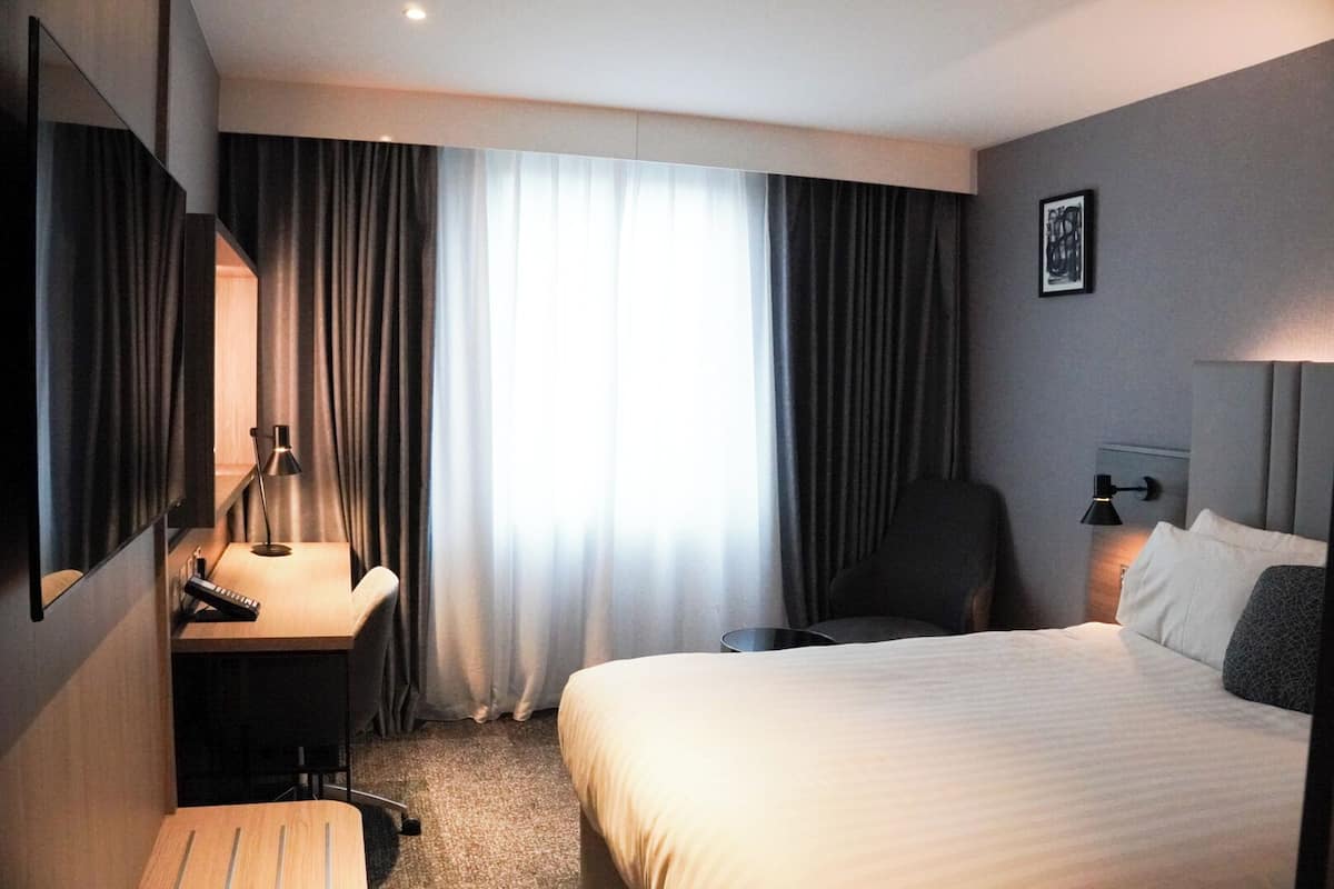 Een hotelkamer met een bed, een bureau met lamp, een stoel en een groot raam met gordijnen.