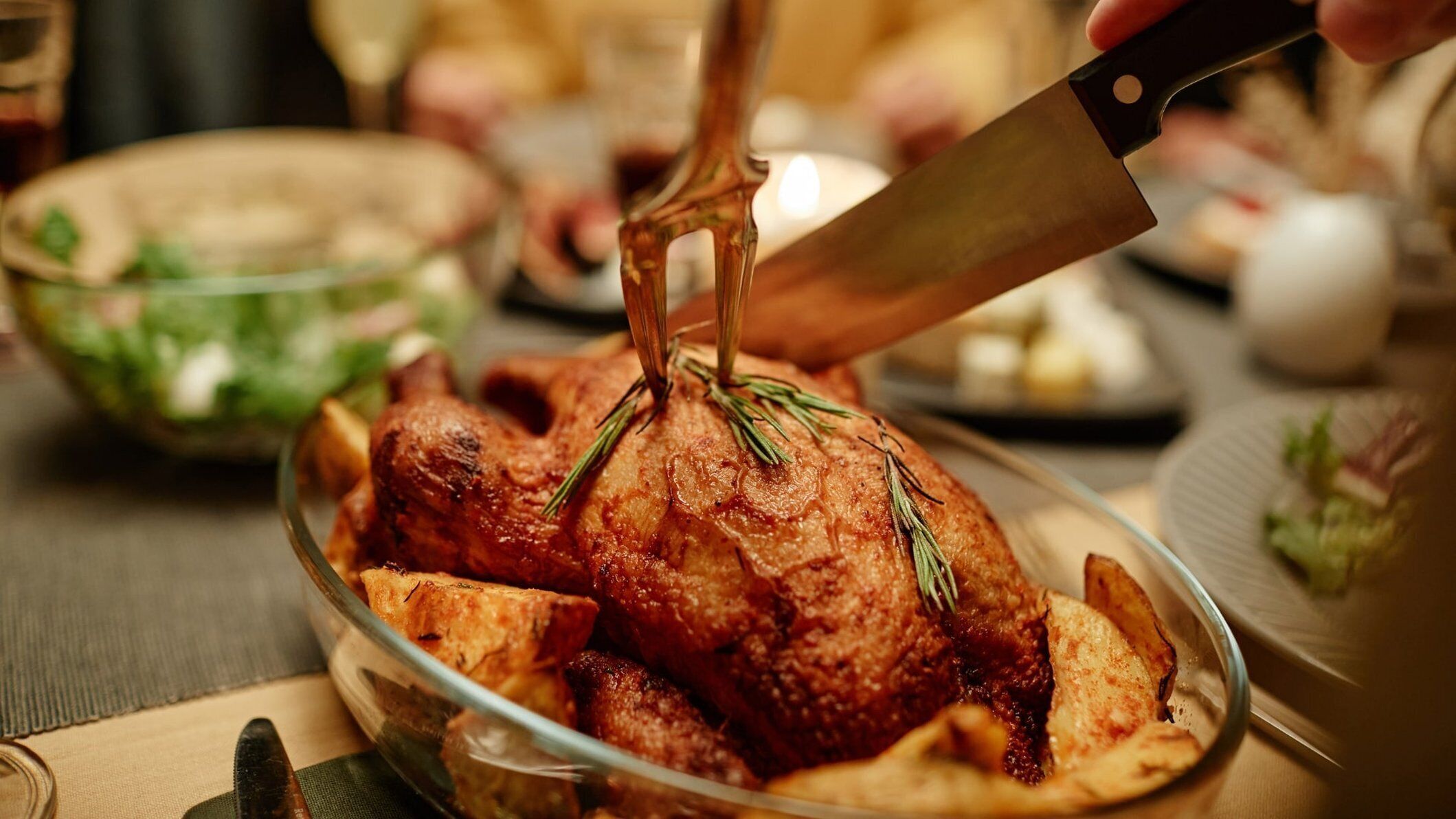 Un pavo asado sobre un plato de cristal con ramitas de romero, rodeado de otros platos y un cuchillo.