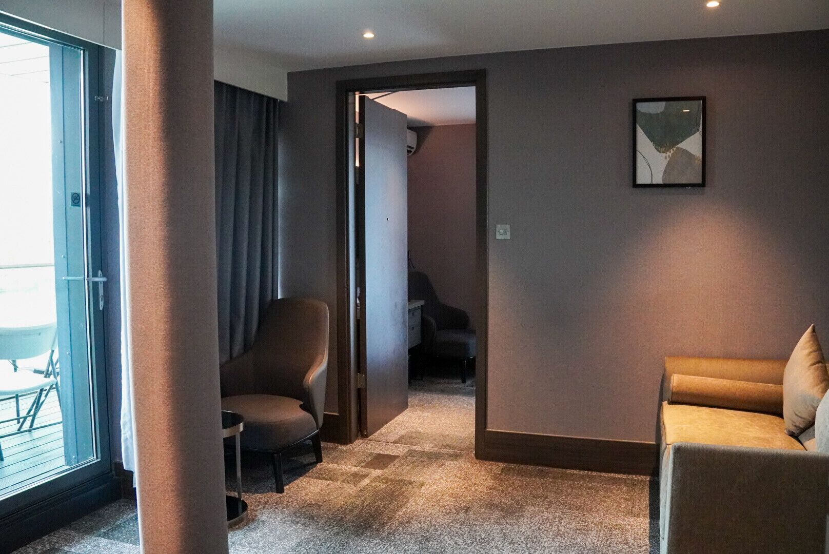 Habitación de hotel con un ventanal grande, una silla, un sofá y una puerta que da a otra habitación.