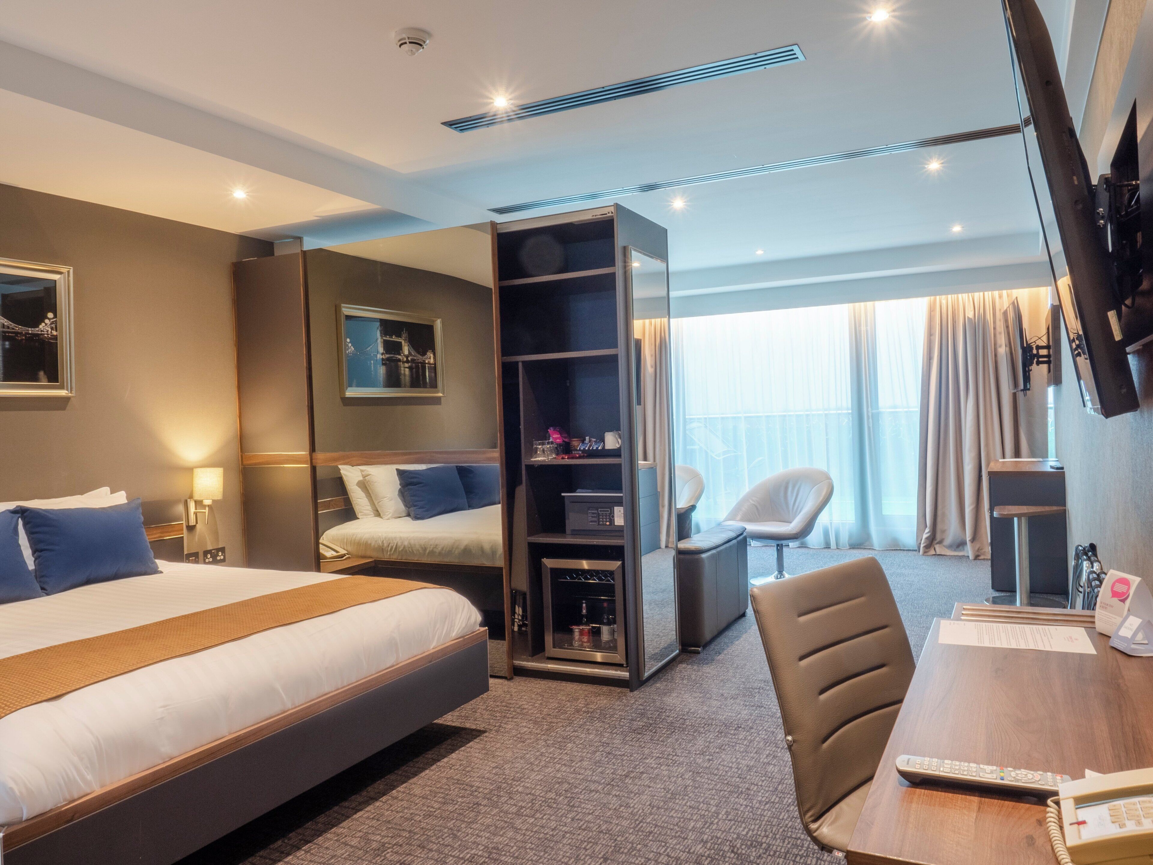 A hotel room with a bed, a desk with a chair, a TV, and a minibar.