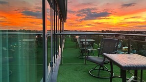 A balcony with green artificial turf, metal chairs, and tables, overlooking a sunset with orange and pink hues.