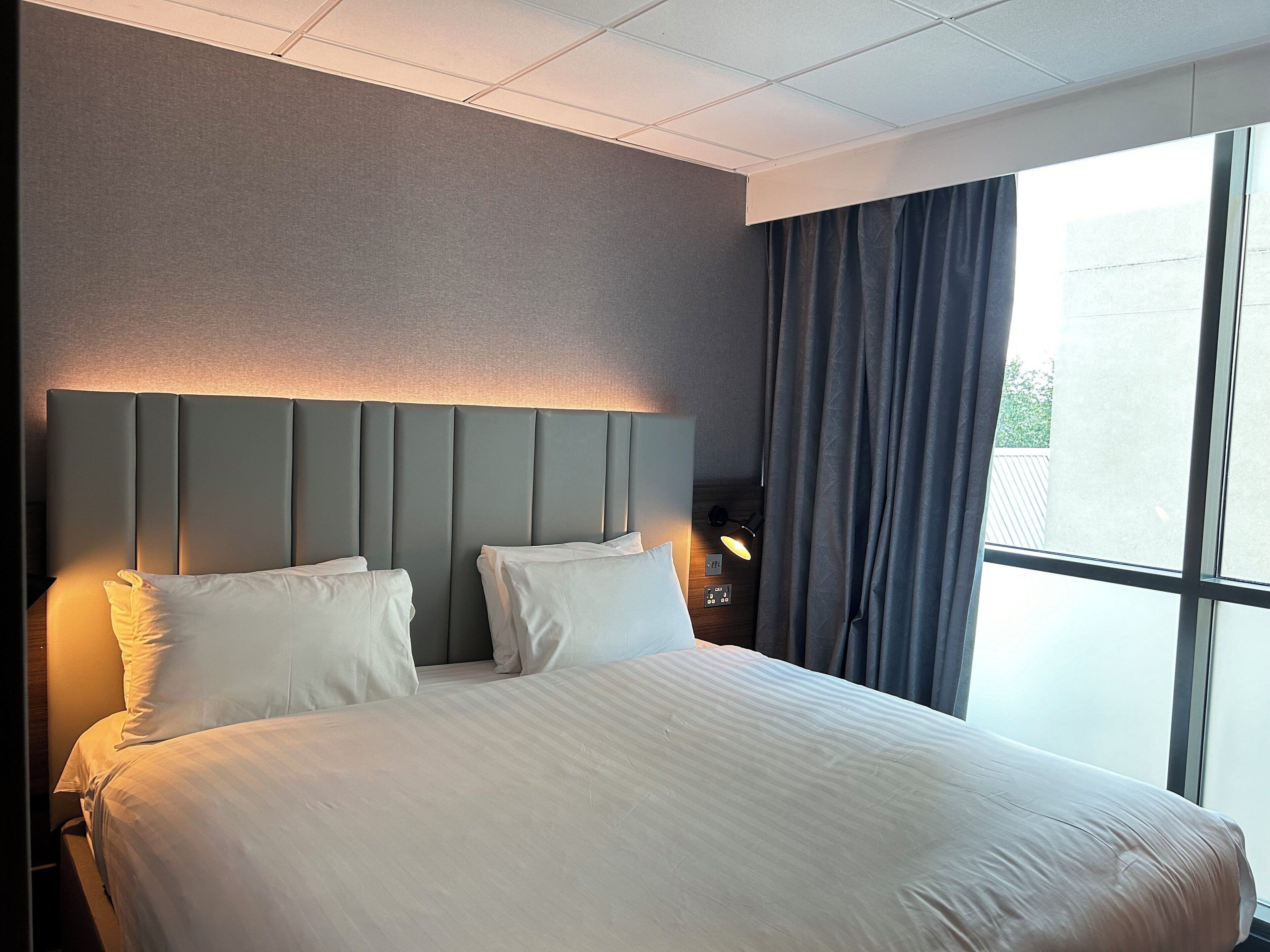 A hotel room with a large bed, a headboard with integrated lighting, a nightstand, a lamp, and a view of the outdoors through a window with curtains.