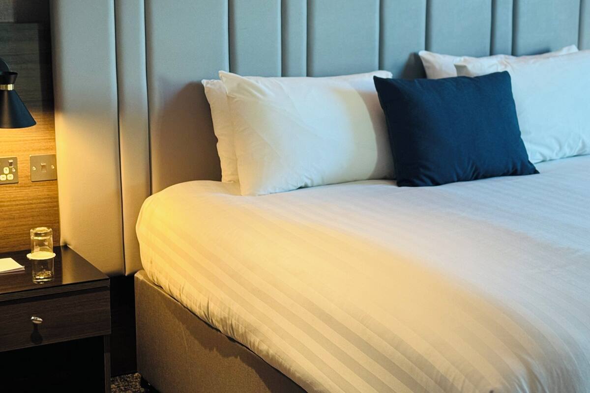Una cama bien hecha con almohadas blancas y azul oscuro, una mesita de noche de madera con una lámpara, y una pared con diseño de paneles verticales.
