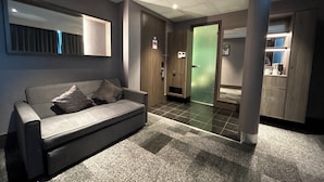 A modern hotel lobby with a grey sofa, a mirror, a wooden door, and a green door leading to a room.