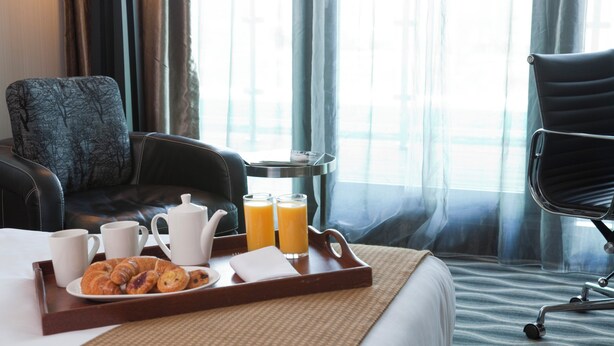 A hotel room with a bed, a dining table with breakfast items, a chair, and a view of the outdoors.
