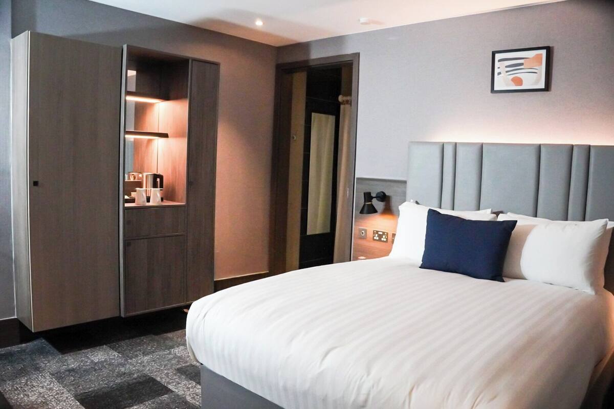 Habitación de hotel con cama, armario, baño y mesita de noche con lámpara.