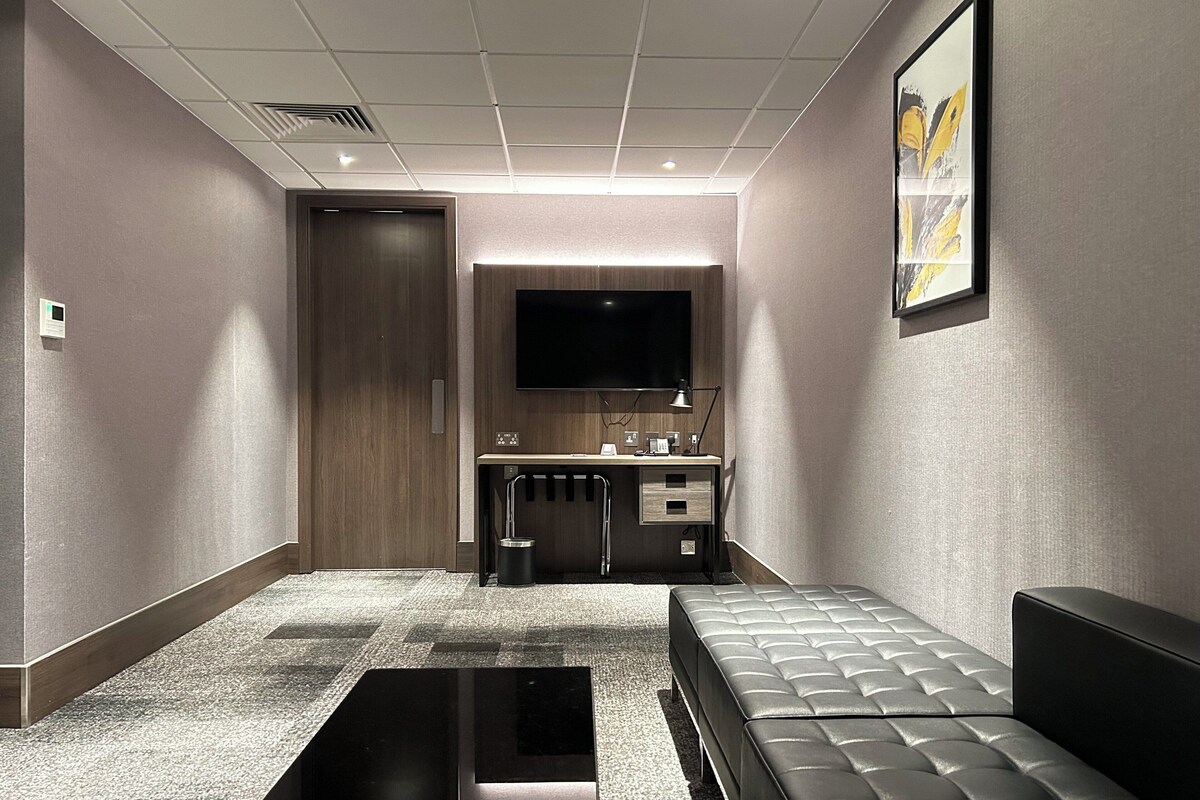 Ein modernes Hotelzimmer mit einem großen Bett, einem Schreibtisch mit Flachbildfernseher, einer kleinen Küchenzeile und einem Badezimmer.