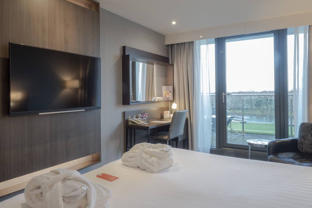 Een hotelkamer met een groot bed, een bureau met stoel, een aan de muur bevestigde televisie en uitzicht op een meer door het raam.