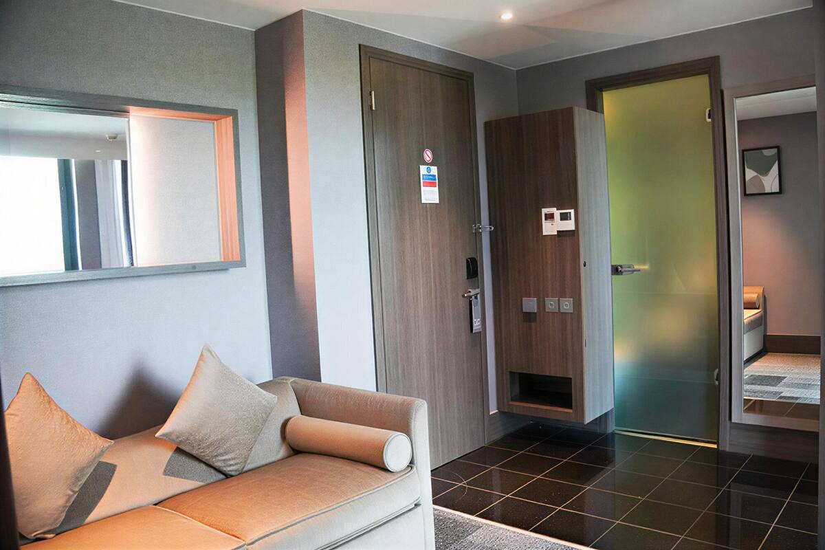 Een moderne hotelkamer met een bank, een spiegel, een houten garderobe, een deur met een geëtste glazen ruit en een klein bed.