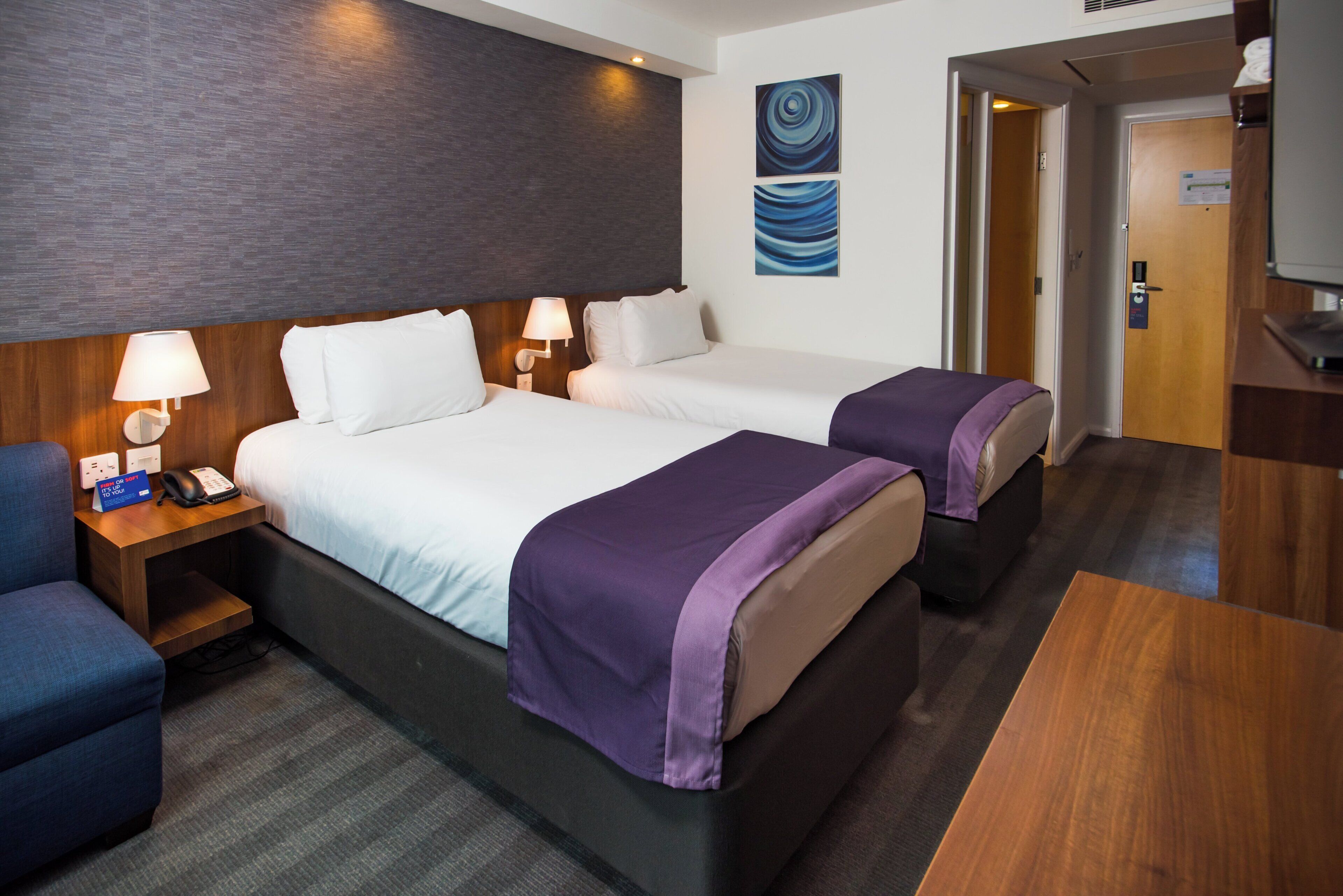 A hotel room with two beds, a desk, a chair, a lamp, and a painting on the wall.