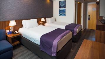 Quarto de hotel com duas camas, uma escrivaninha, uma cadeira, um abajur e um quadro na parede.
