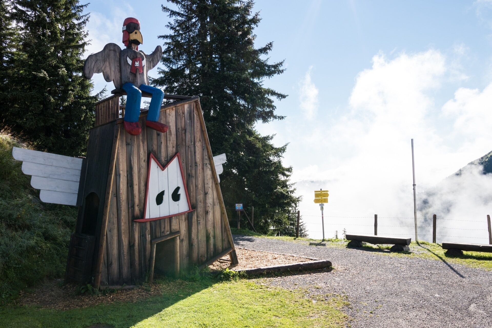 A wooden structure with a figure resembling a bird, a bench, and a signpost in a natural setting.