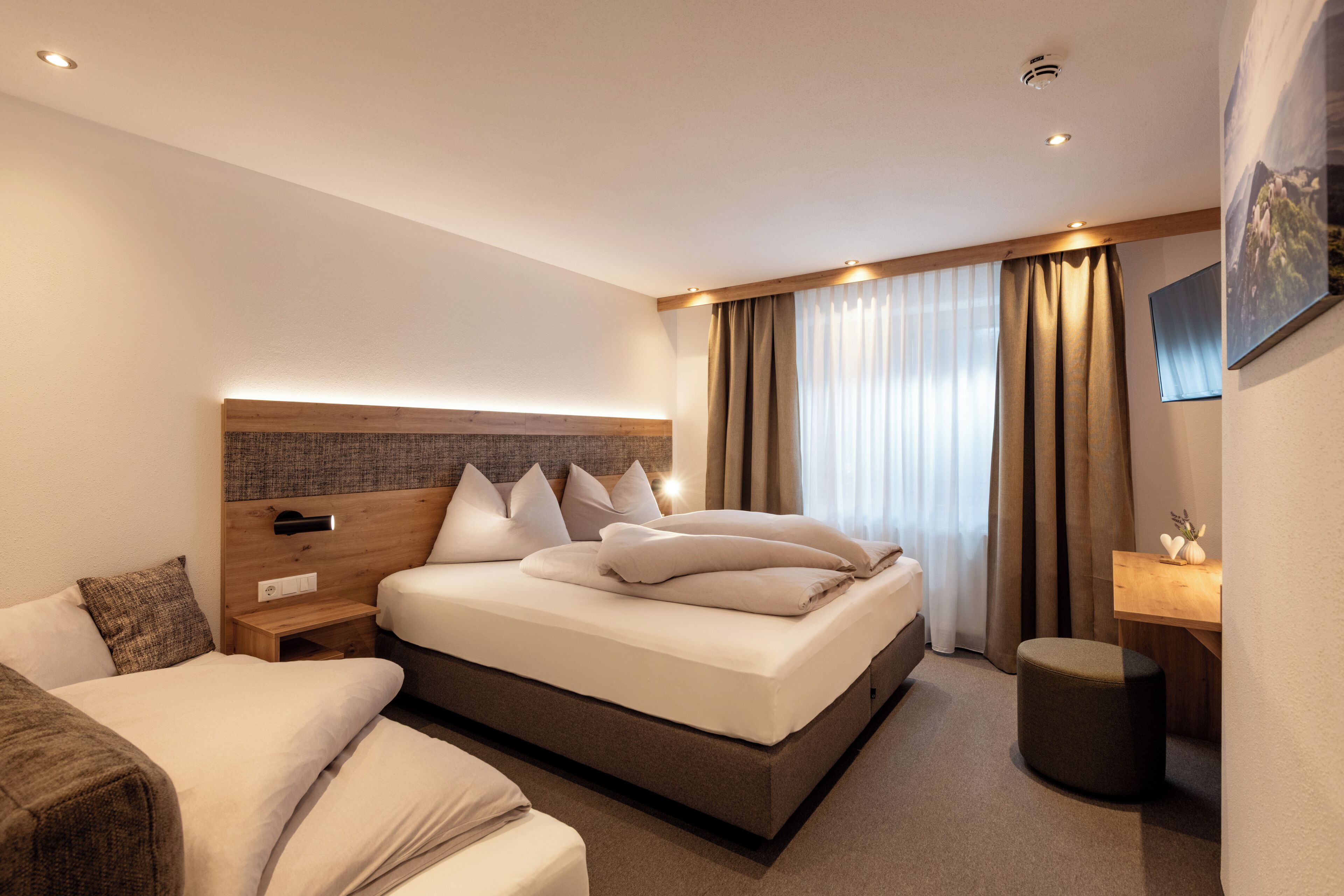 A modern hotel room with a large bed, a bedside table, a desk, and a small ottoman.