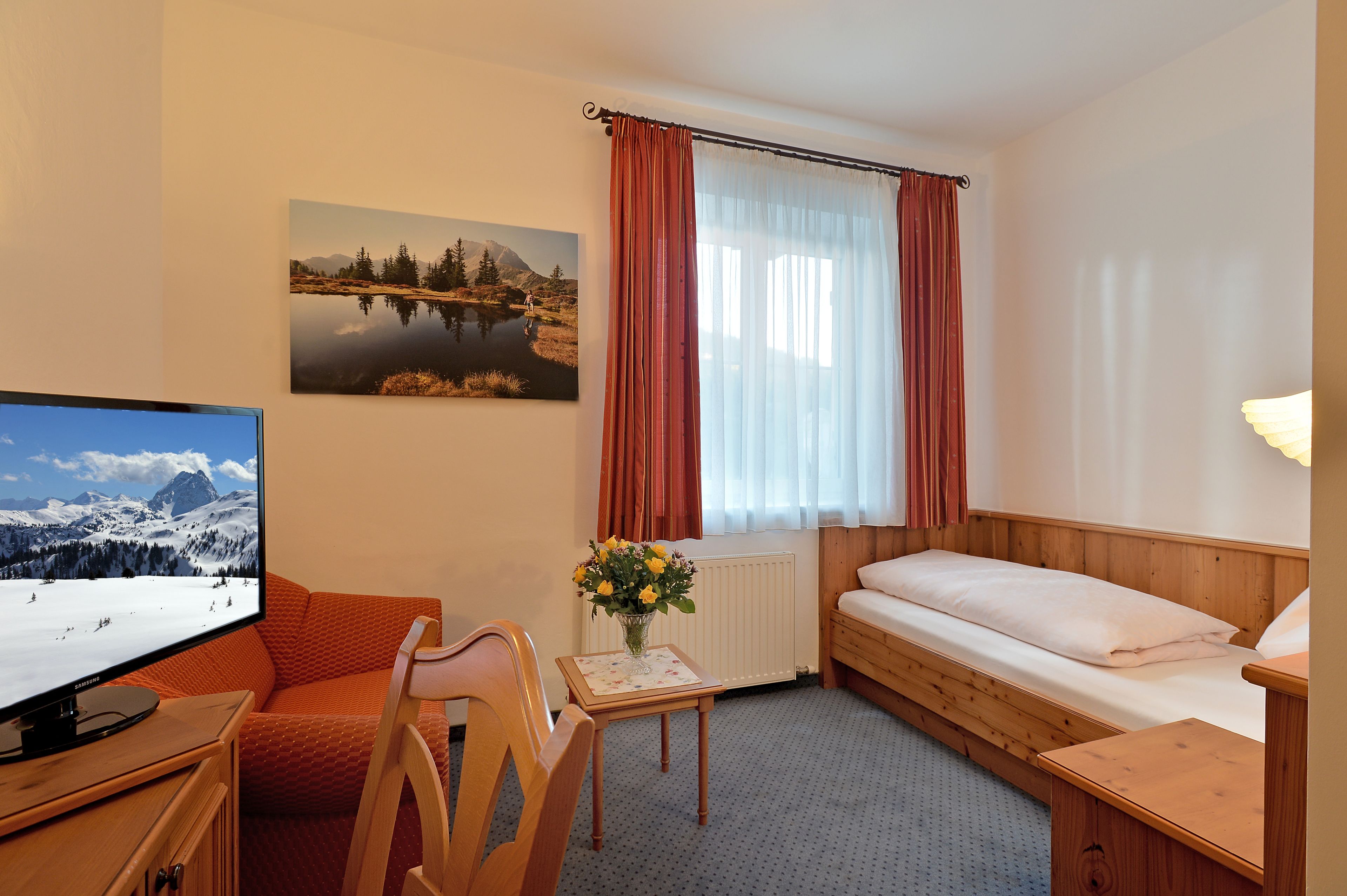 Quarto de hotel com cama, escrivaninha com computador, cadeira, mesa com um vaso de flores, janela com cortinas e um quadro na parede.