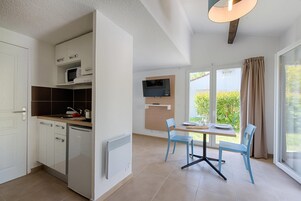 A modern kitchen with white cabinets, a built-in microwave, and a dishwasher. There is a dining area with blue chairs and a table, and a TV mounted on the wall.