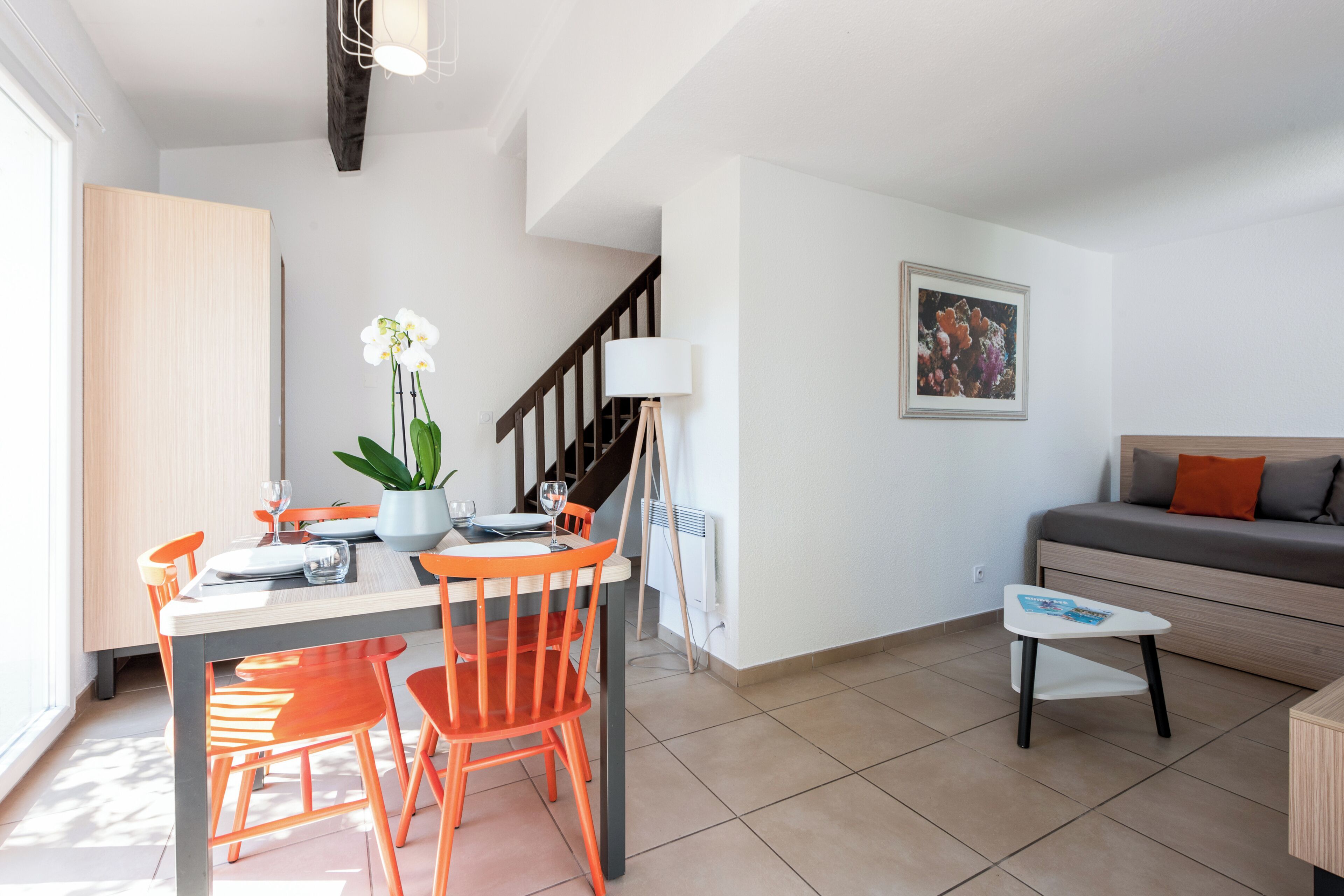 A modern living room with a dining area, featuring a staircase, a sofa, a small table, and a potted plant.