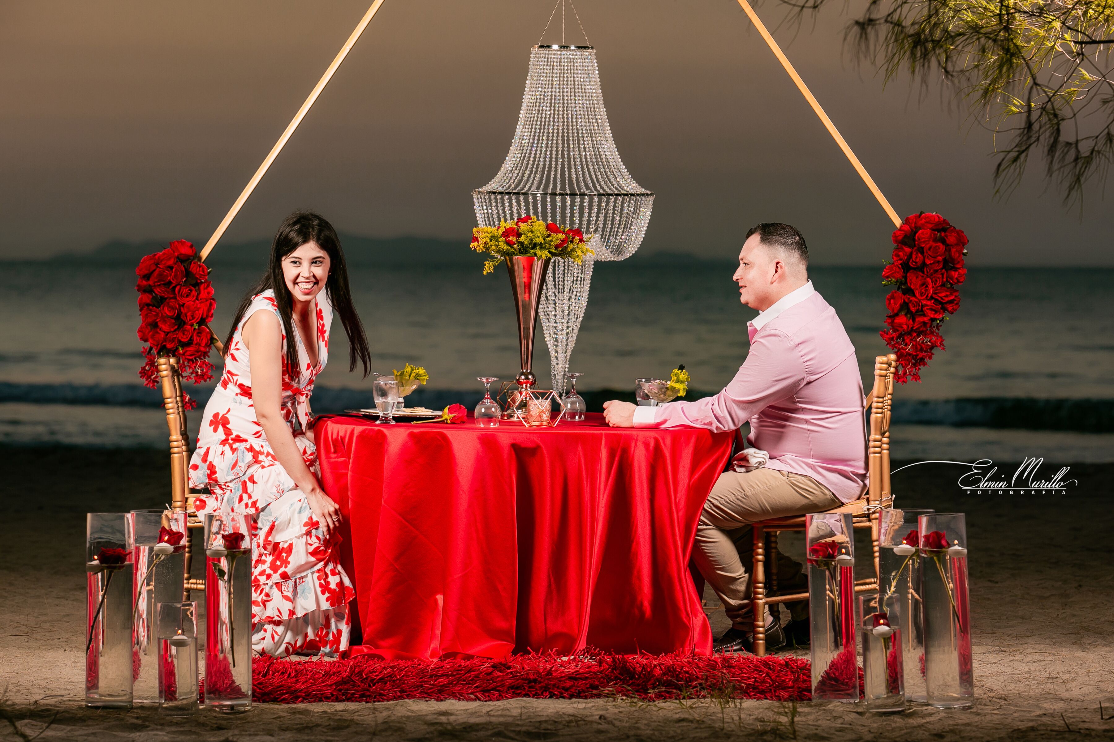 Una pareja sentada a una mesa con mantel rojo, rodeada de jarrones de vidrio con flores, en una playa con vista al mar.