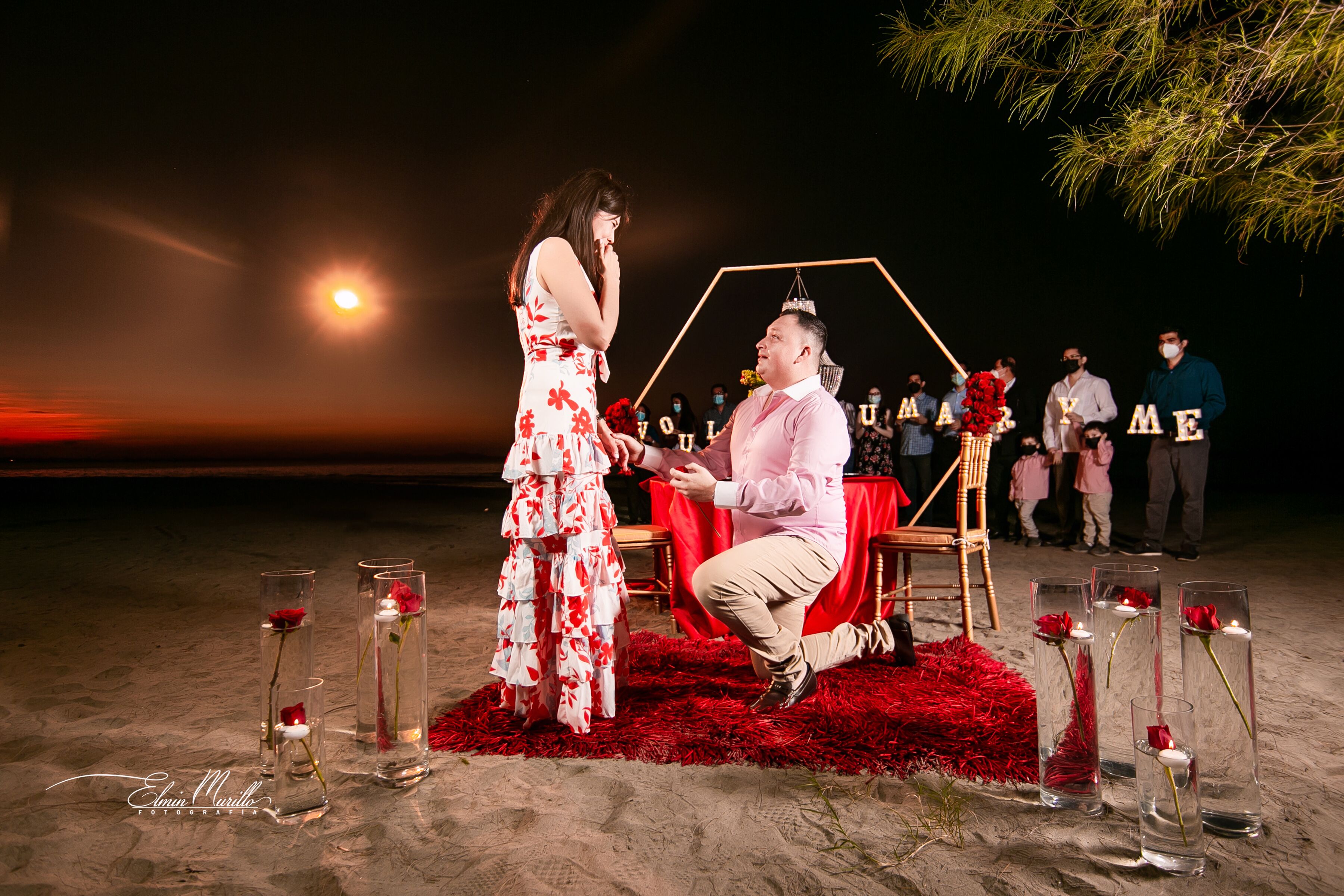 Escena en la playa con un hombre proponiendo matrimonio a una mujer, rodeados de invitados y una mesa decorada con velas y flores.