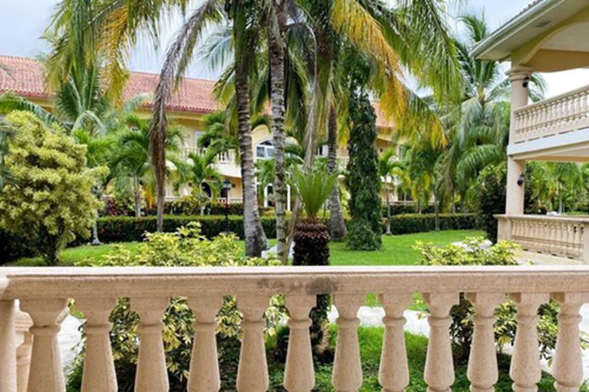 A balcony with a view of a well-maintained garden, palm trees, and a building with a red-tiled roof.