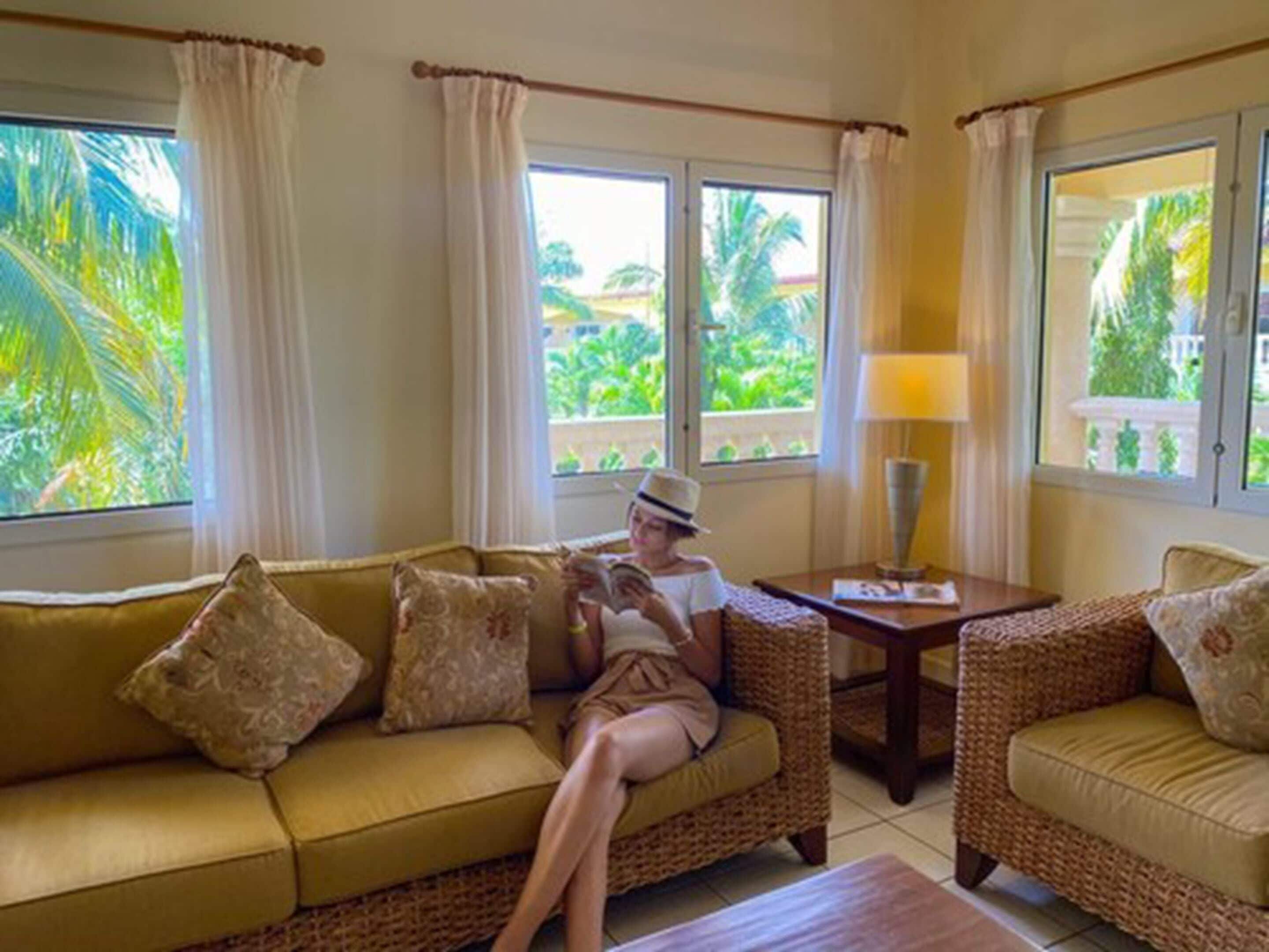 A room with a sofa and two wicker chairs, a table with a lamp, and large windows with curtains.