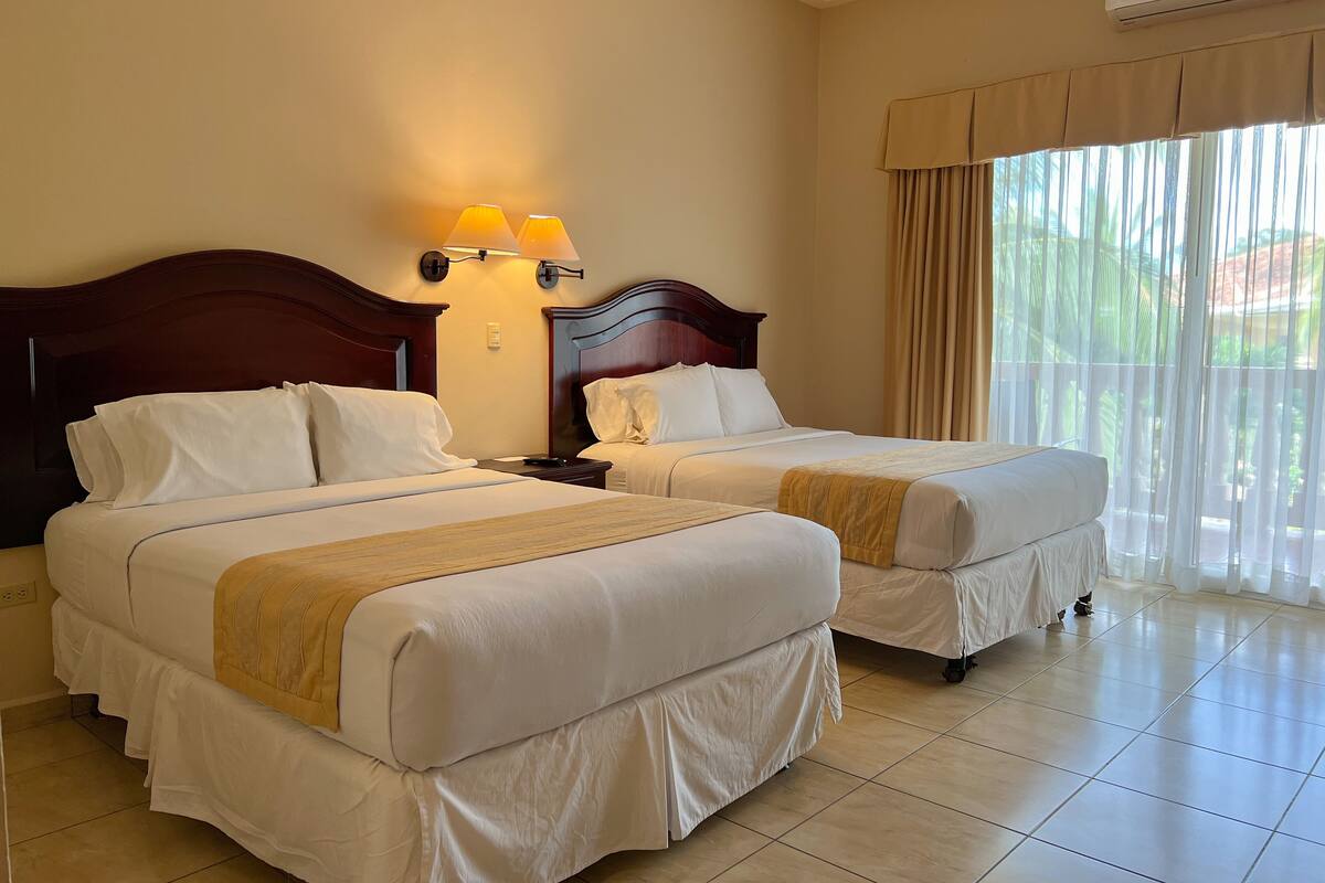 A hotel room with two beds, a wooden headboard, a nightstand with a lamp, and a large window with curtains.