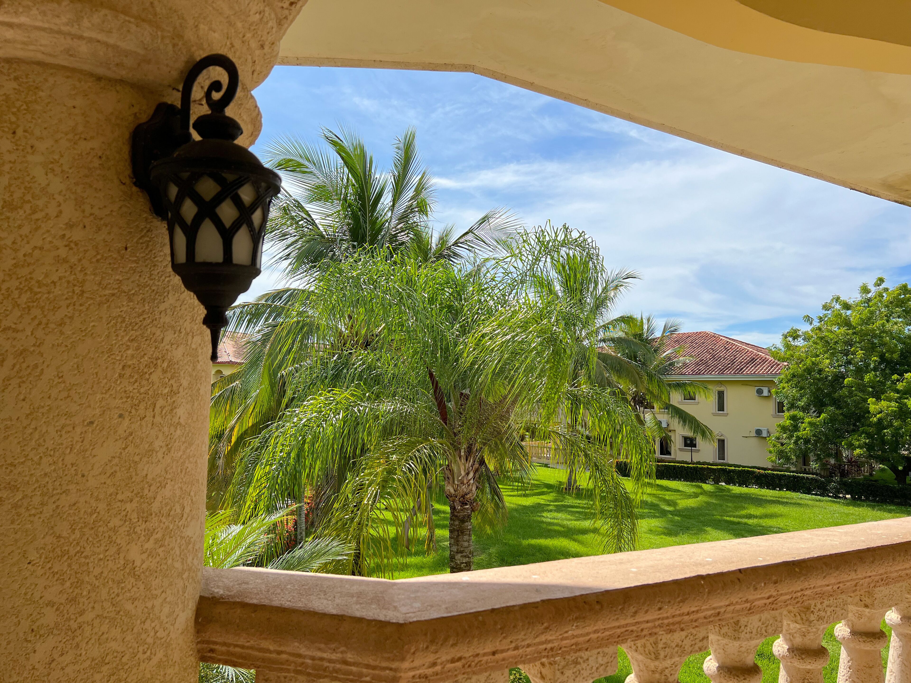 A balcony with a stone balustrade, a wall-mounted lantern, and a view of a well-maintained lawn with palm trees and a building with a red-tiled roof.