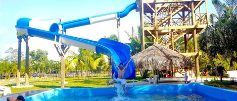 Parque acuático con un tobogán de agua azul, una piscina y una estructura de techo de paja.