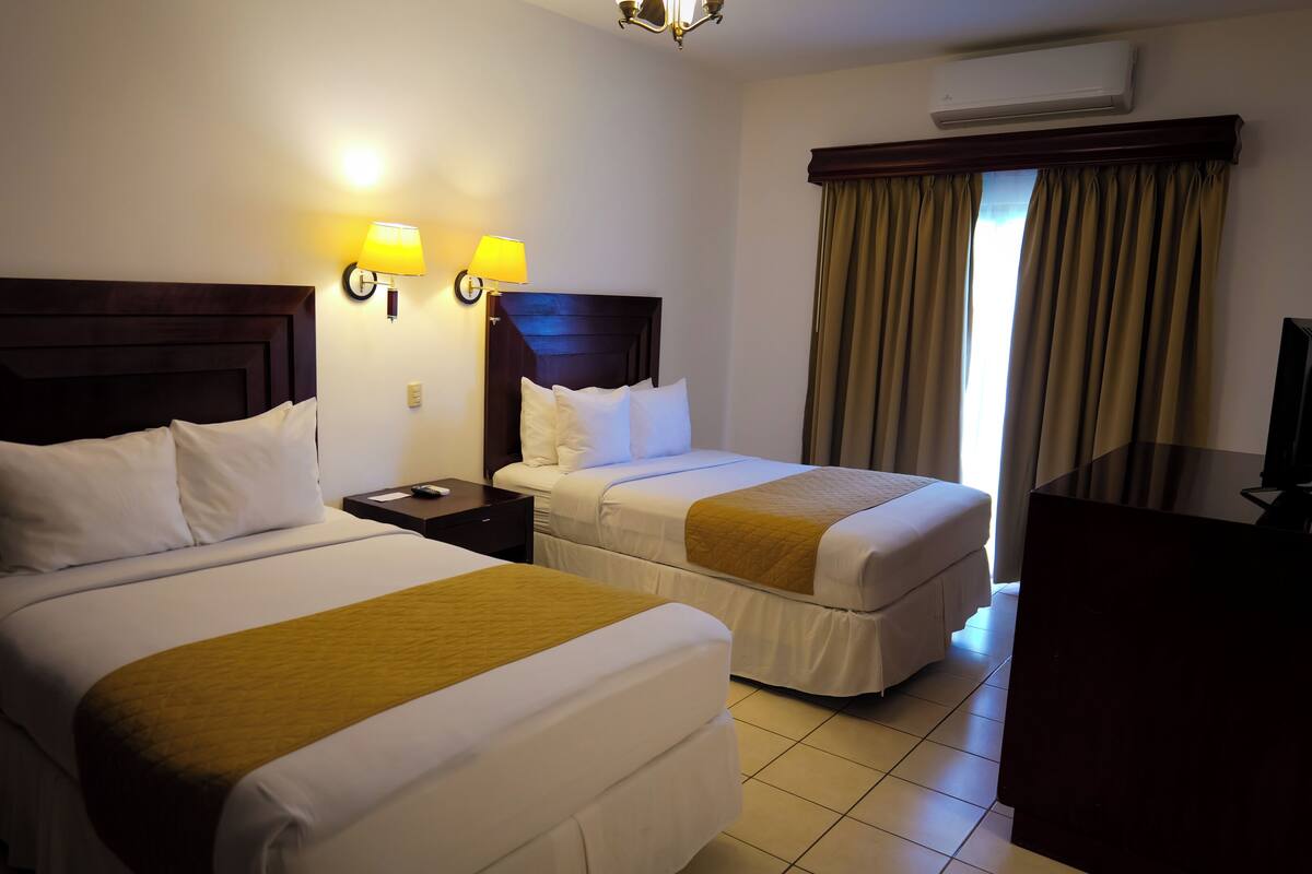 A hotel room with two beds, a desk, a TV, and a window with curtains.