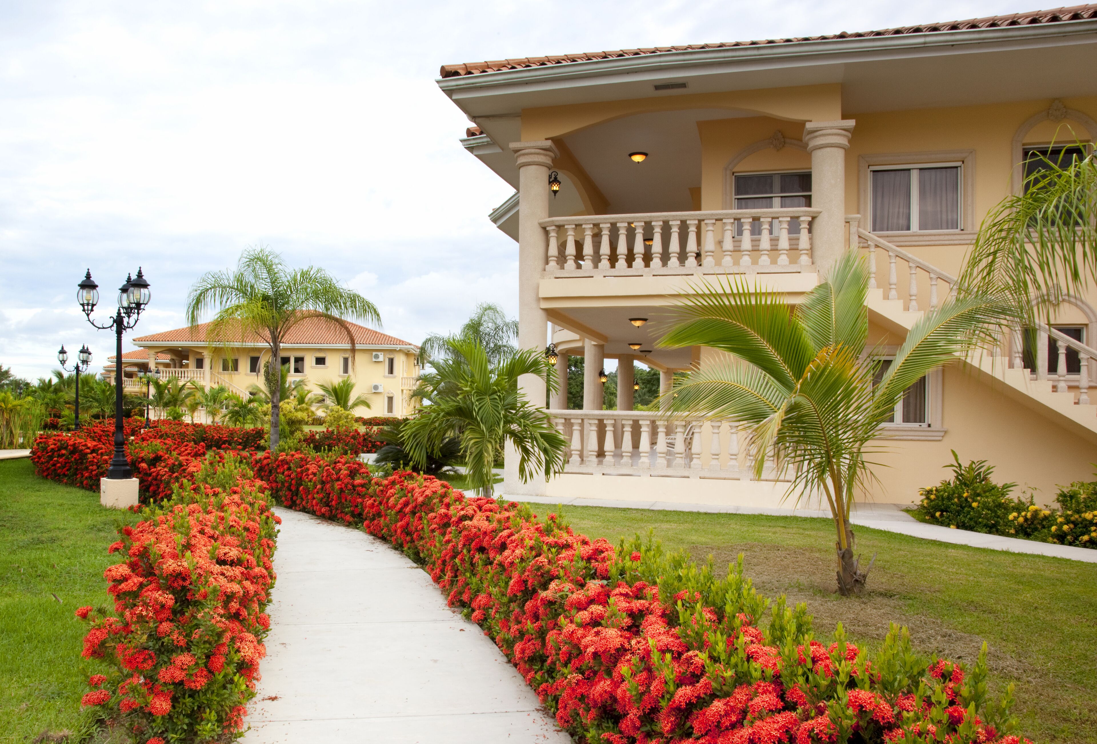 Un edificio grande de dos pisos con un balcón y un jardín bien cuidado con flores rojas y palmeras.