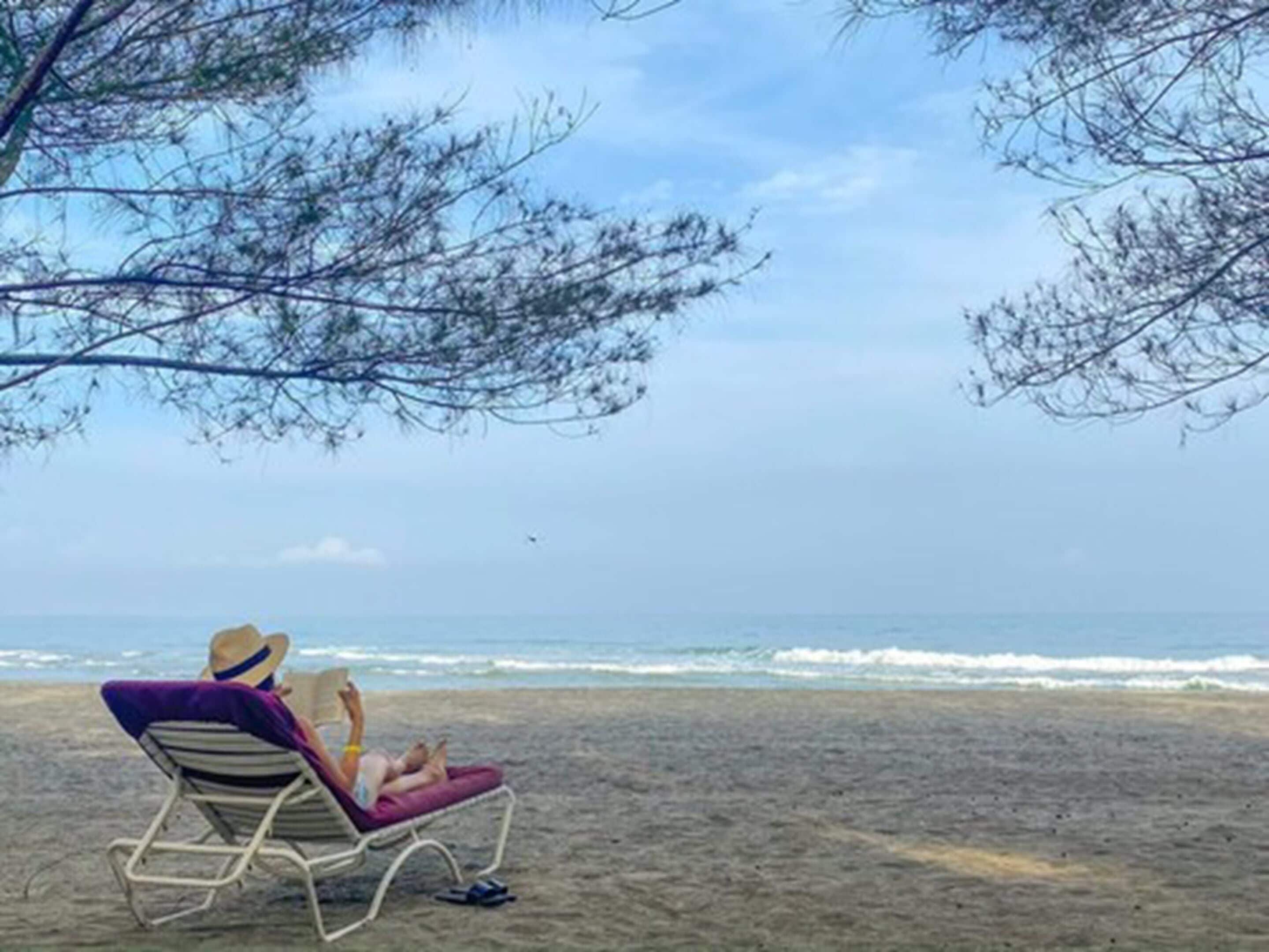Une scène de plage où l’on voit une personne se détendre sur une chaise longue, à l’ombre d’un arbre.