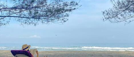 A beach scene with a person relaxing on a beach chair under a tree.