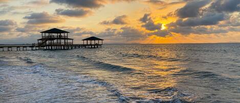 A pier extending into the sea with two covered structures, likely for relaxation or dining, under a sunset sky.