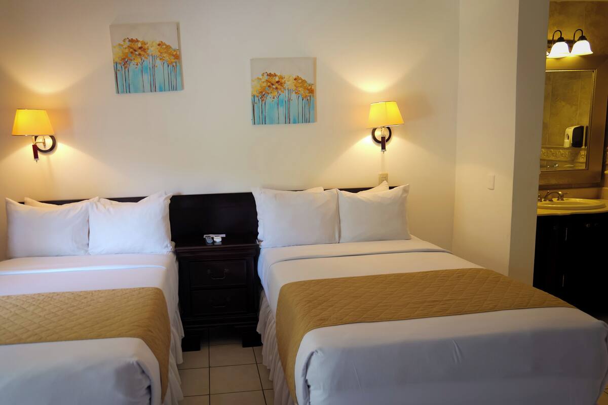 A hotel room with two beds, a nightstand, and a bathroom with a sink and mirror.