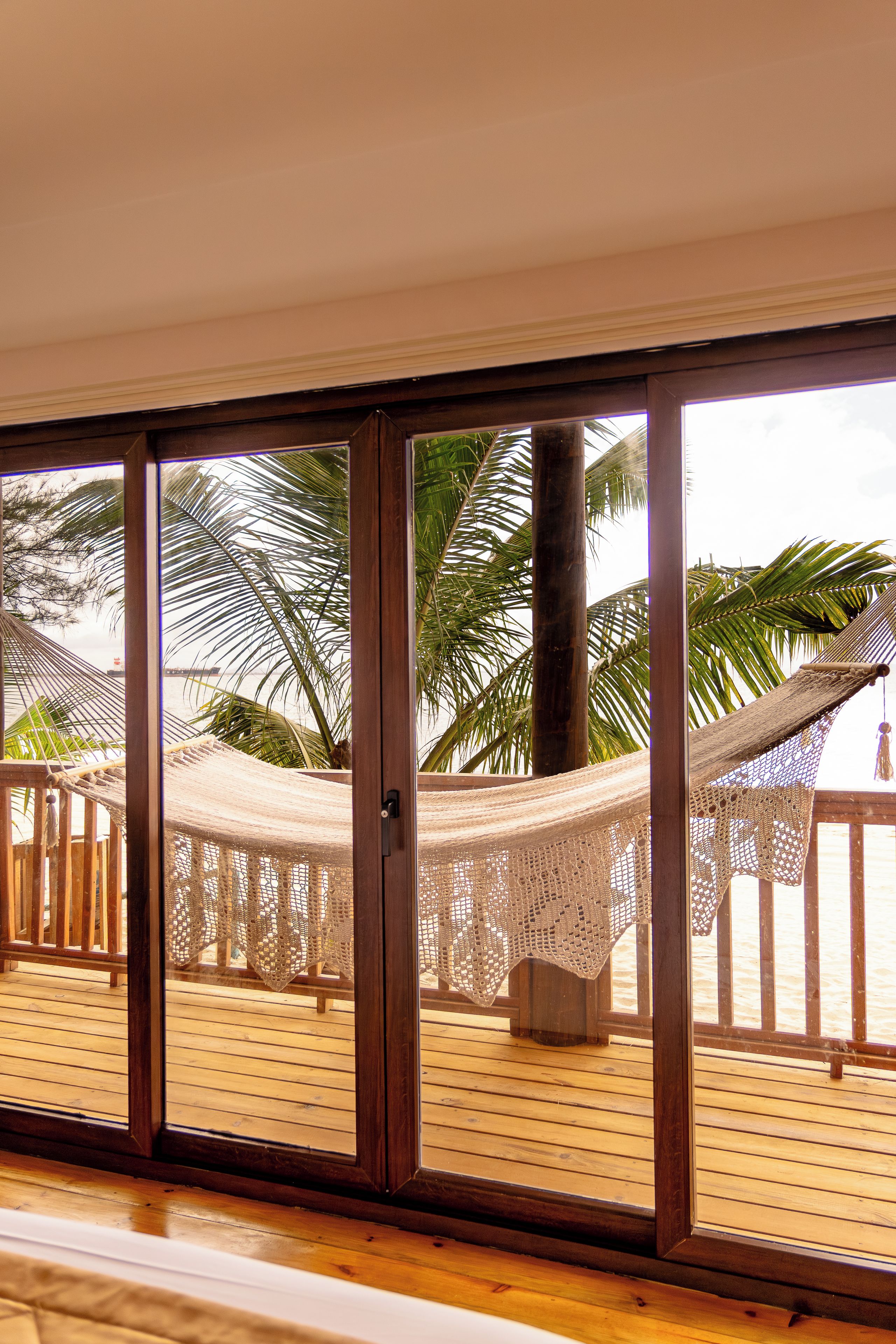 A wooden deck with a hammock and a wooden bench, overlooking a beach with palm trees.