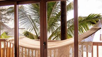 A wooden deck with a hammock and a wooden bench, overlooking a beach with palm trees.