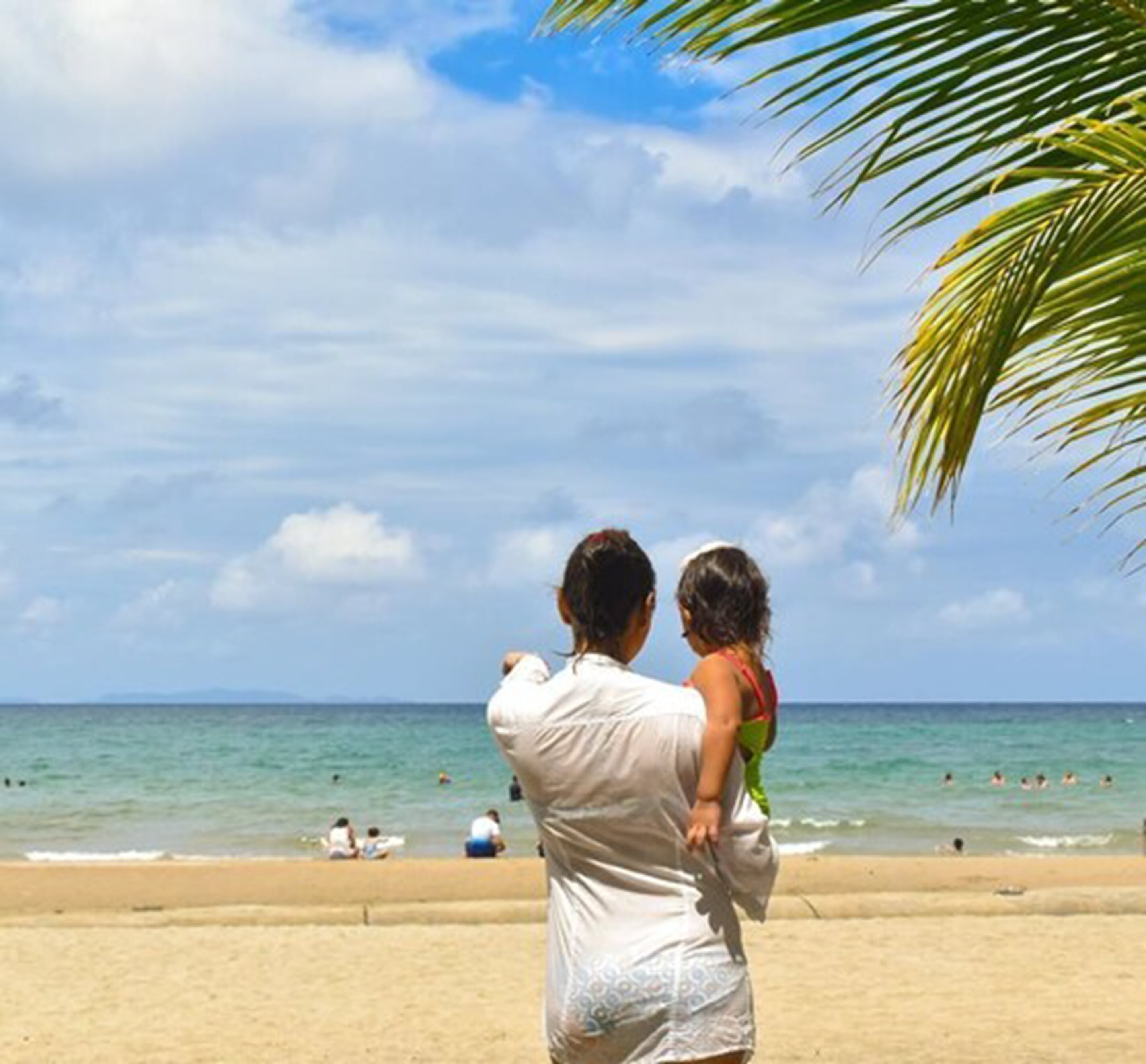 A beach scene with a person holding a child, palm trees, and the ocean.