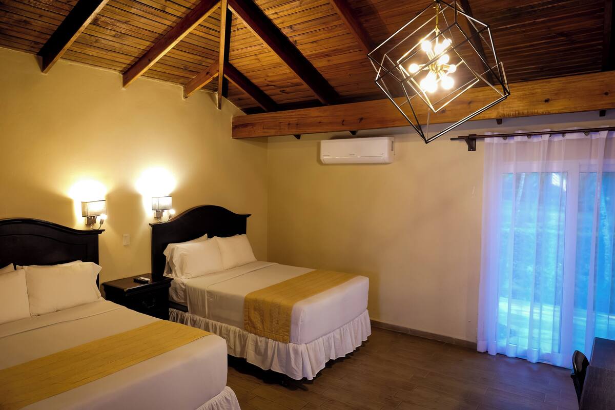 A hotel room with two beds, wooden ceiling beams, a modern chandelier, and a window with sheer curtains.