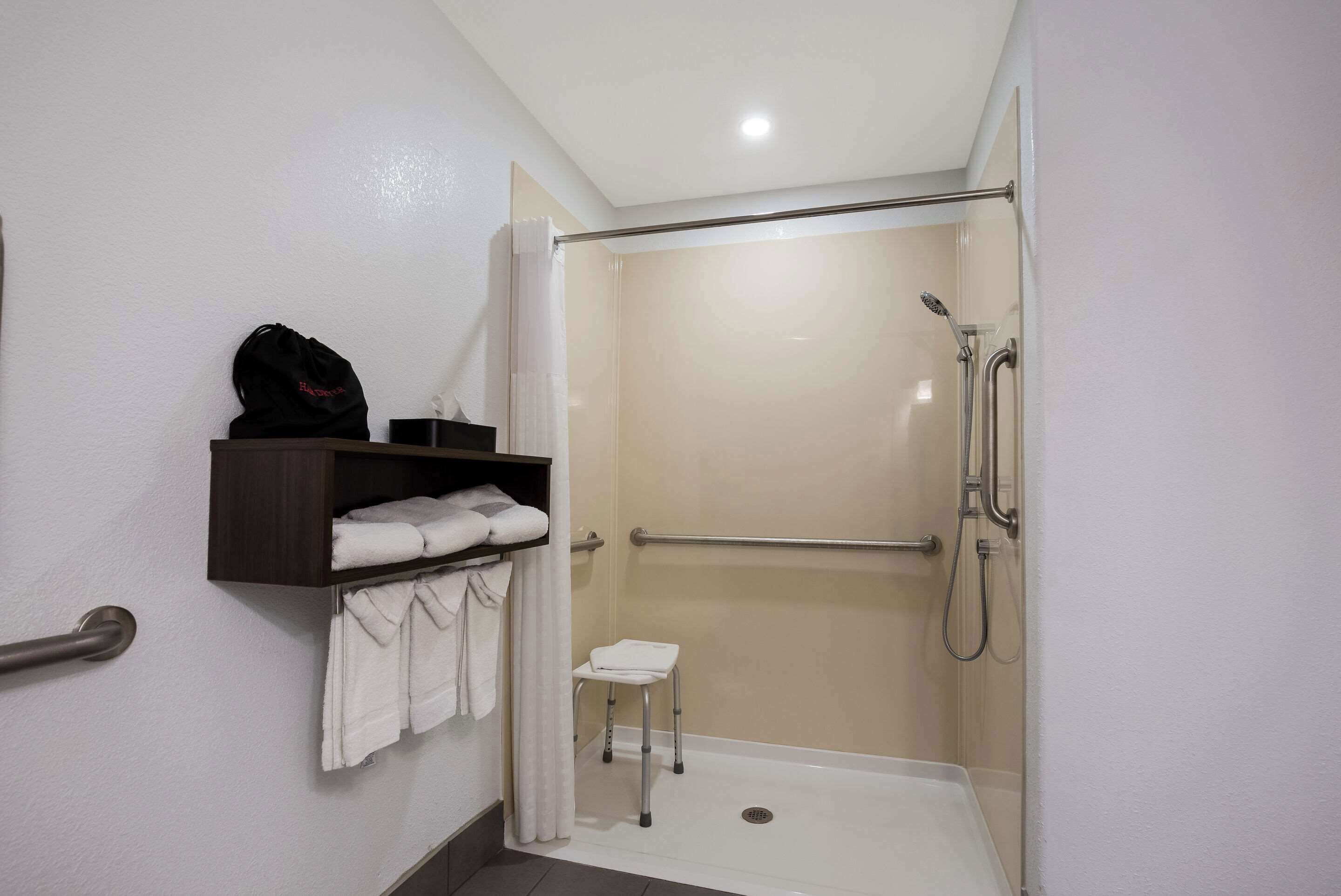 A bathroom with a shower, a small stool, a shelf with towels, and a grab bar.