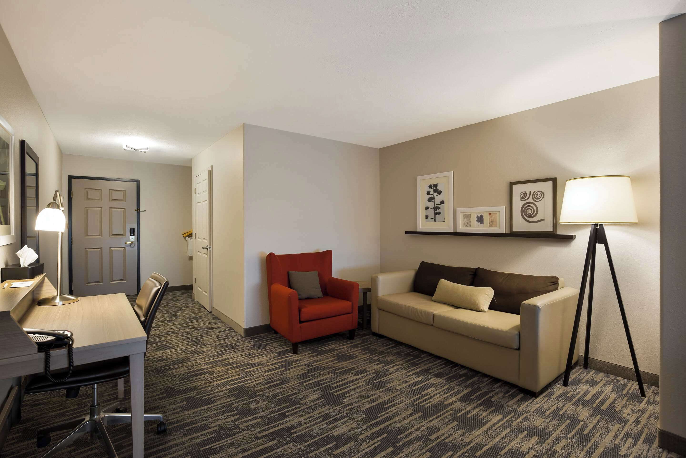 A hotel room with a desk, chair, sofa, and a lamp.